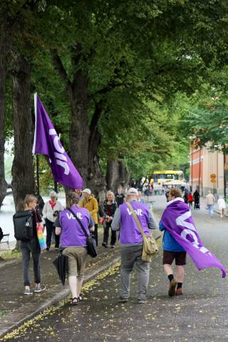 Four people with purple vests and a flag with the Logo of Volt from the back walking along a street under trees